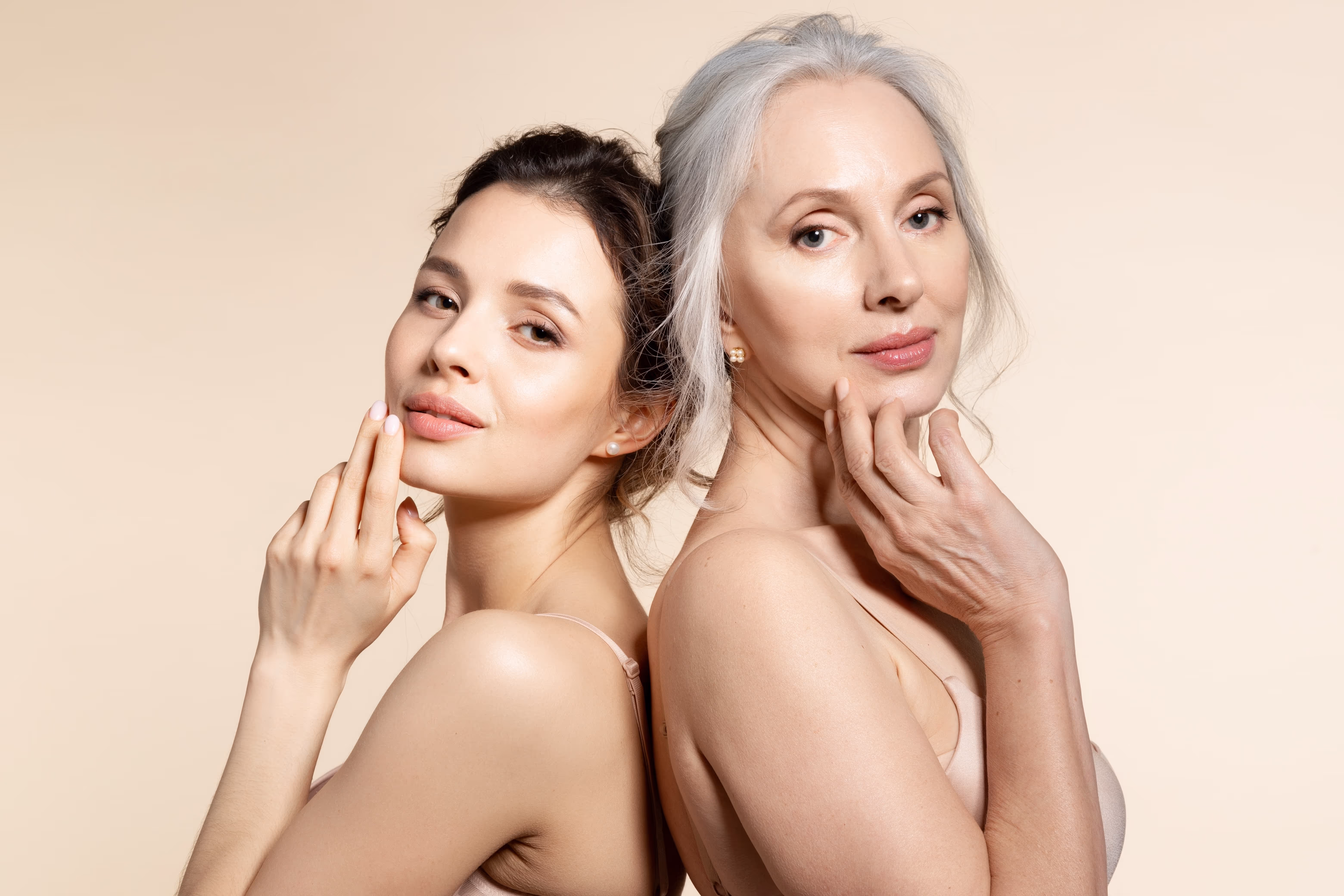 Younger and older woman standing back to back, touching their faces, both with clear skin and neutral expressions.