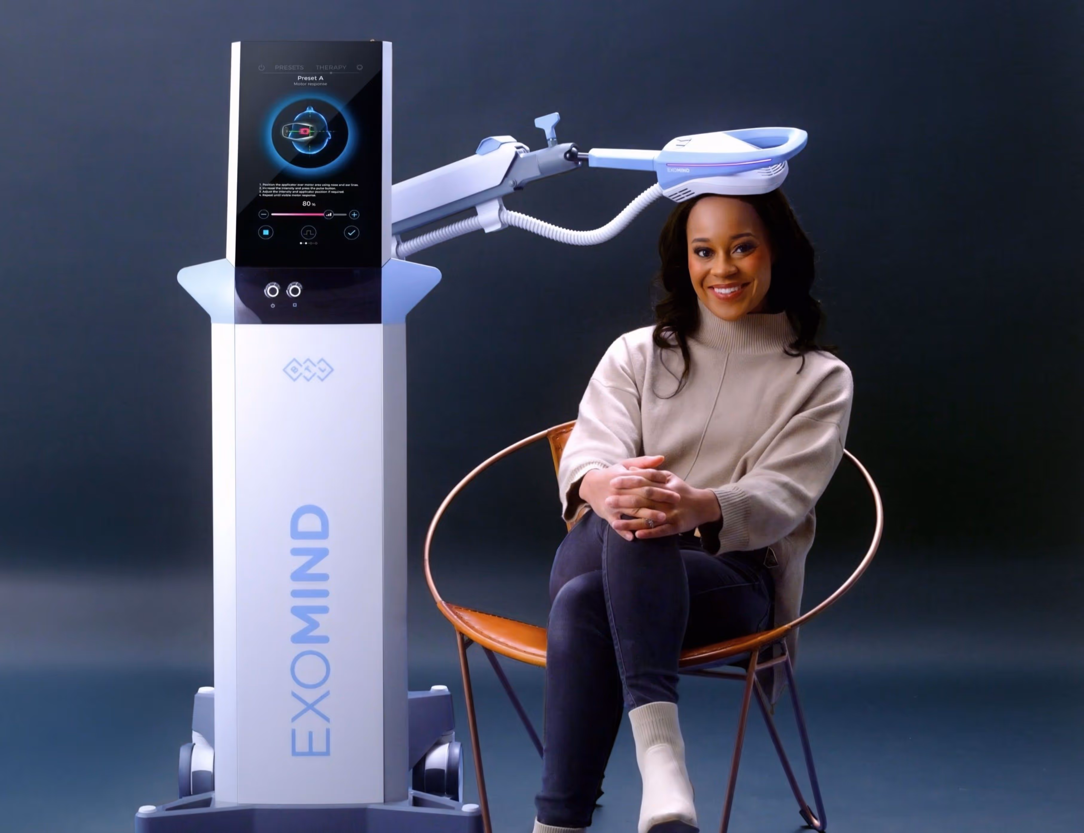 Woman sitting with legs crossed next to an ExoMind medical device with a head applicator.