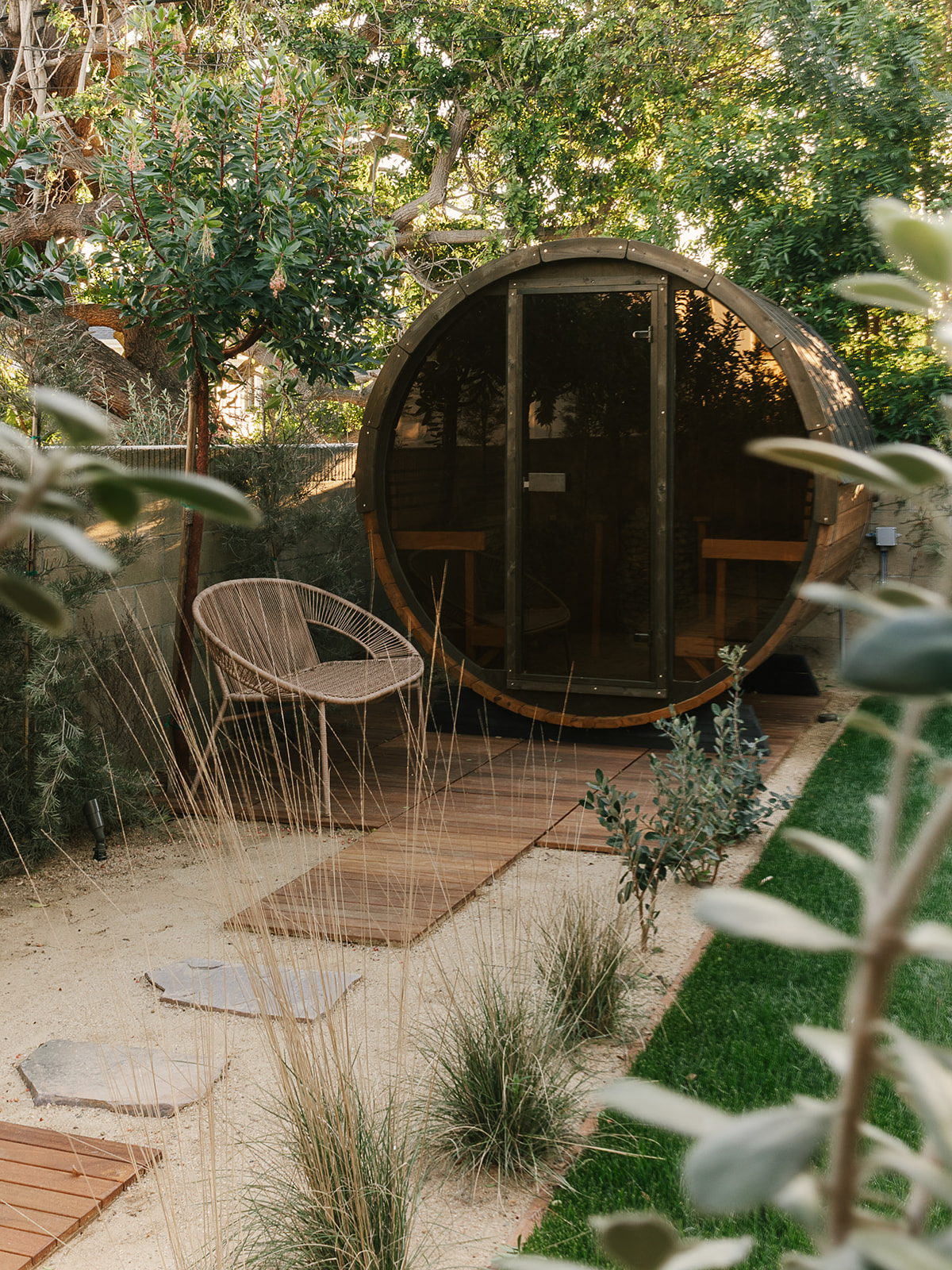 Outdoor wooden barrel sauna with glass door in a garden surrounded by trees, plants, a rattan chair, and a wooden pathway.
