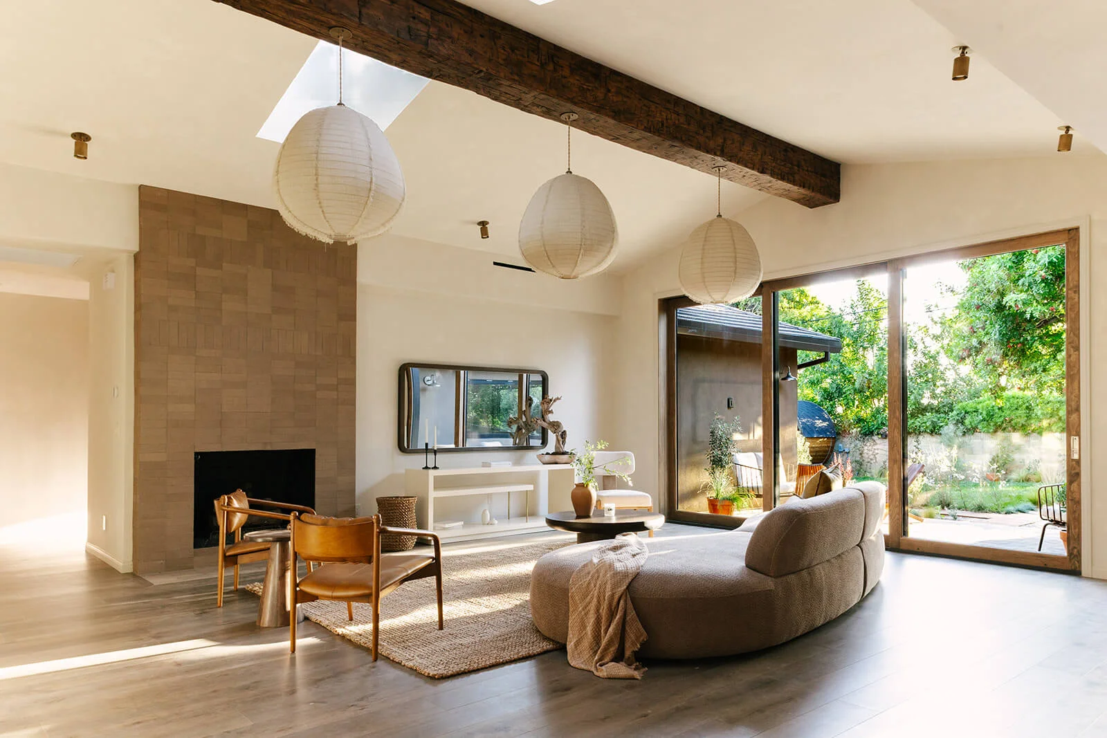 Modern living room with beige curved sofa, leather chairs, a fireplace with a tiled surround, and large sliding glass doors opening to a garden.