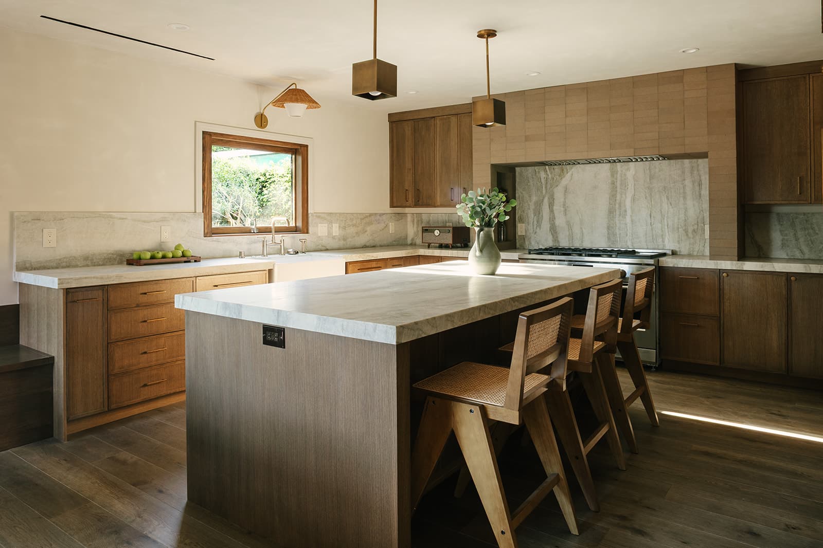 Modern kitchen with large marble island, wooden cabinets, three wooden chairs, a window, and pendant lights.