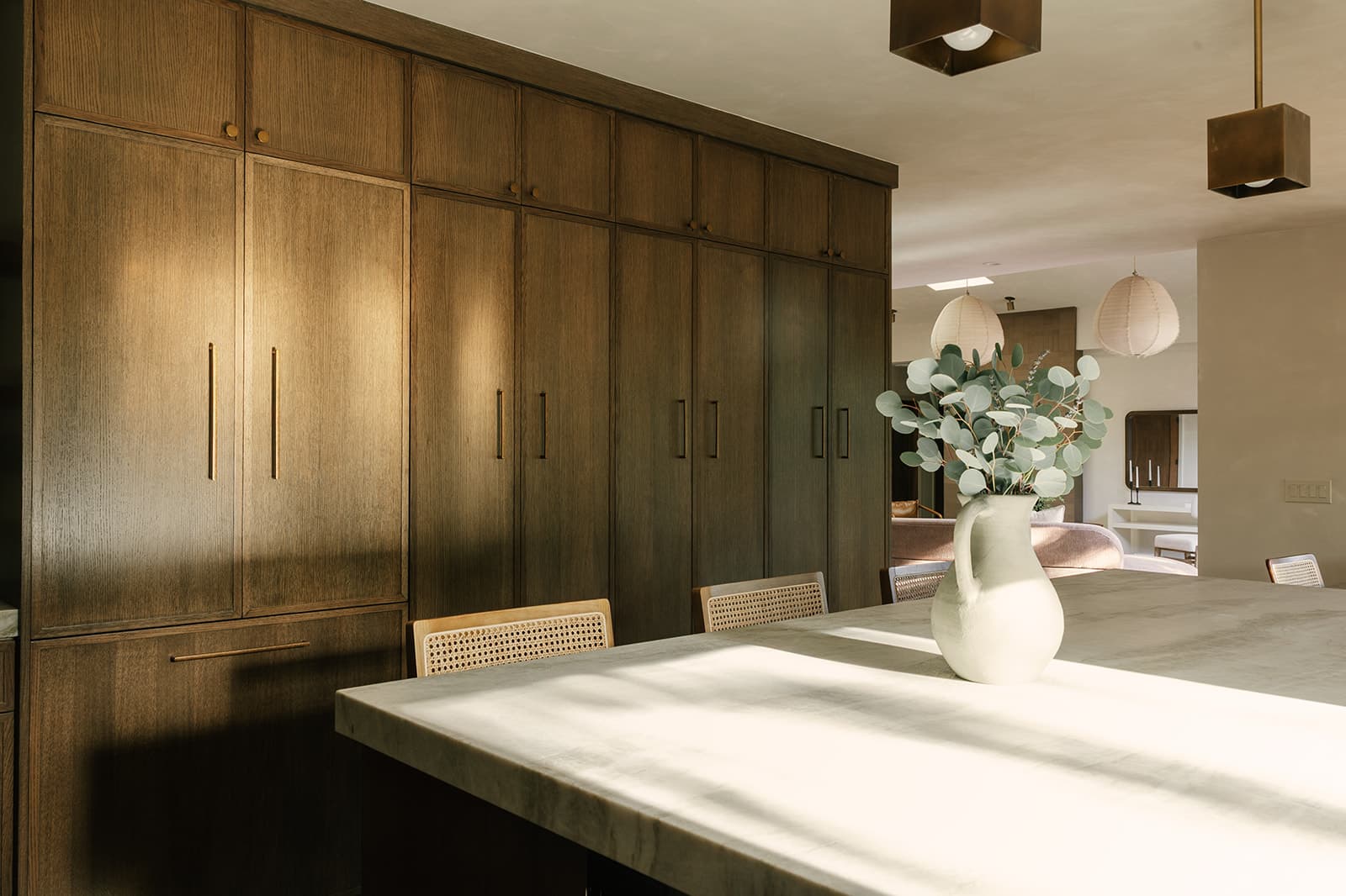 Bright kitchen with large marble island, woven chairs, wooden cabinets, and a white vase with eucalyptus branches.
