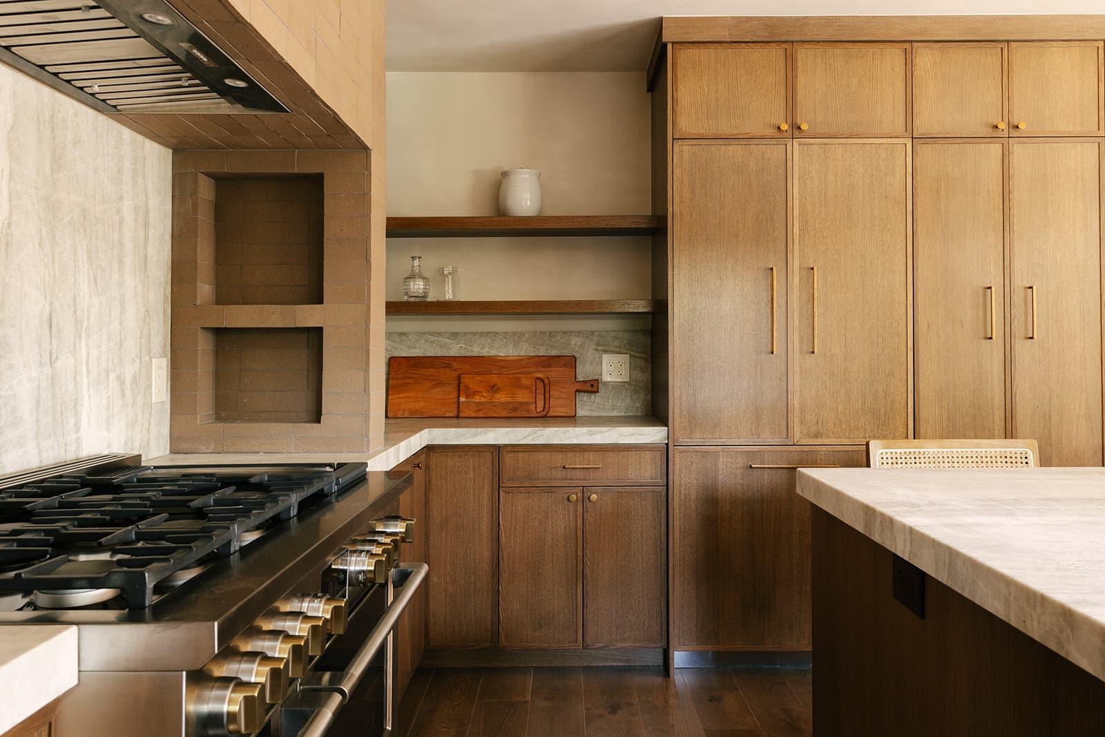 Modern kitchen with wooden cabinetry, marble countertops, a stainless steel stove, and open shelves holding glassware and a ceramic jug.