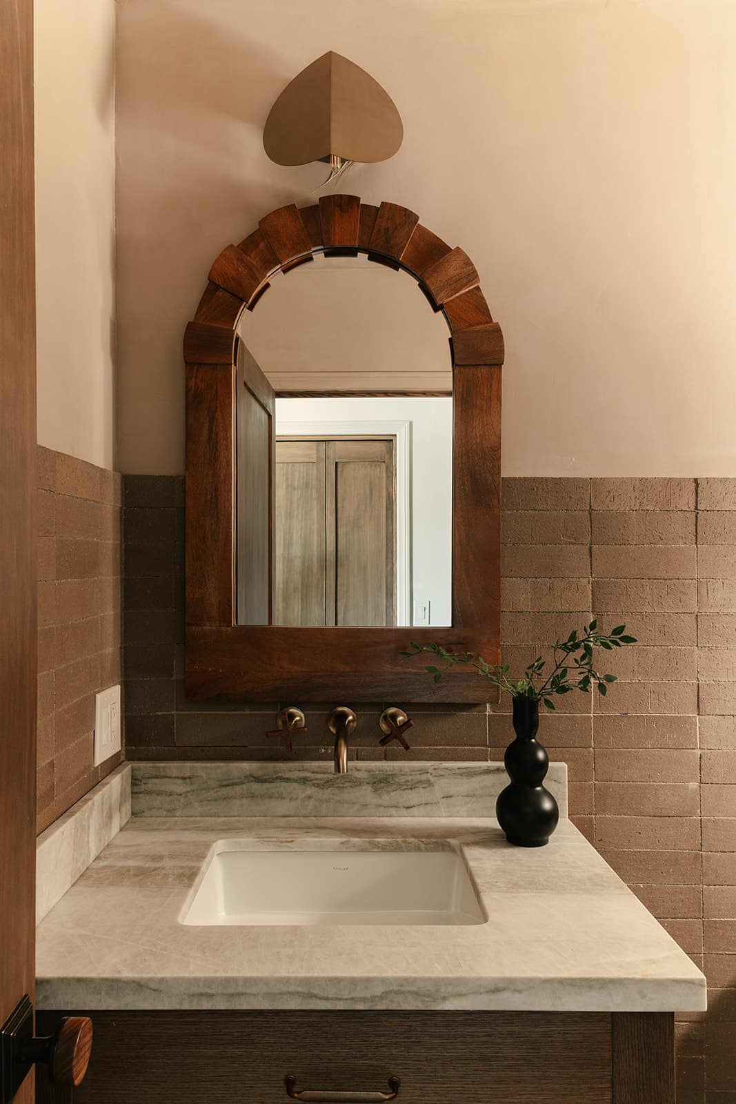 Bathroom vanity with a rectangular marble countertop sink, wall-mounted bronze faucet, wooden arched mirror, and a black vase holding green branches.