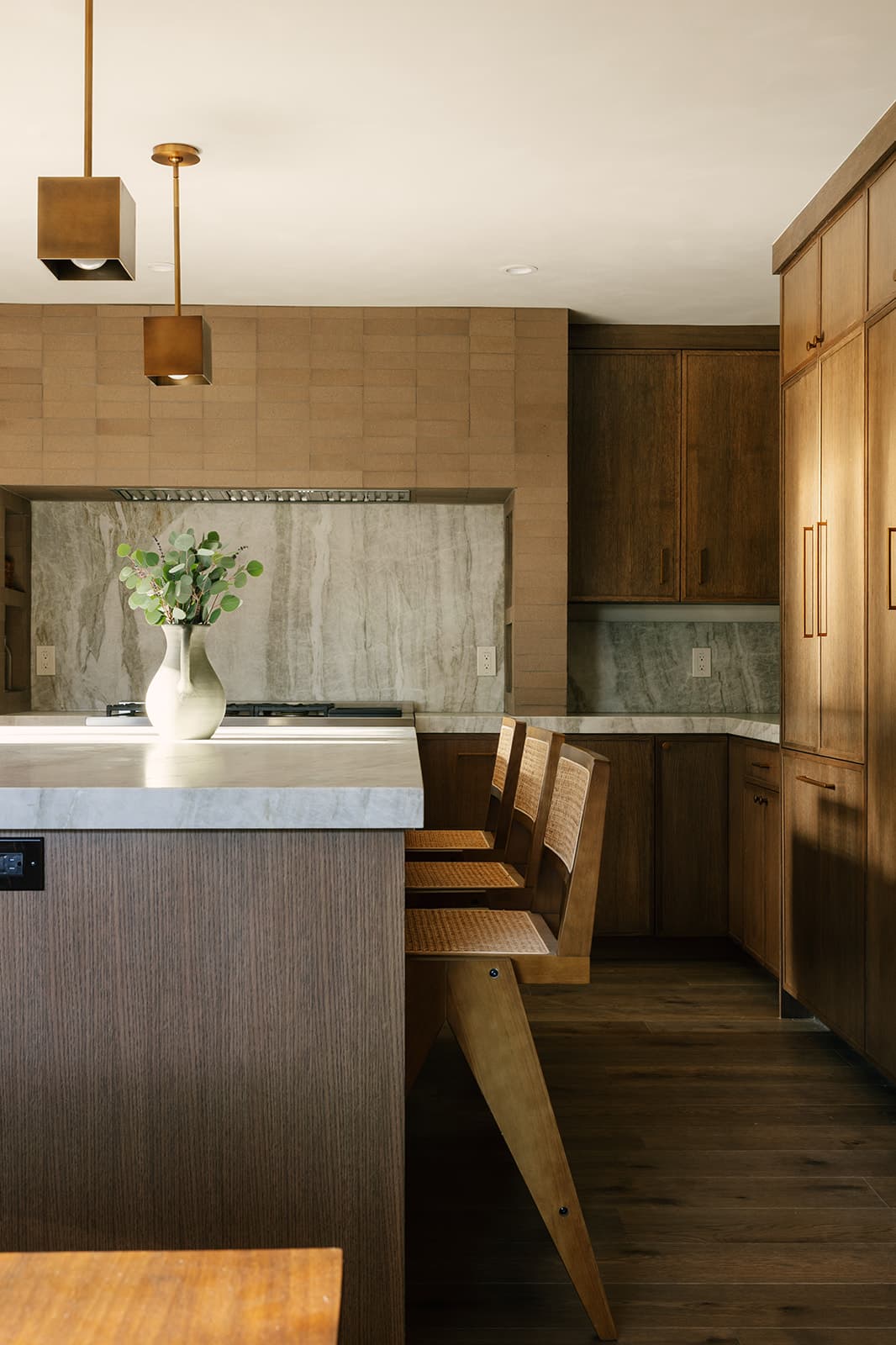 Modern kitchen with wooden cabinets, marble countertop island, two wooden chairs, and a vase with green foliage.