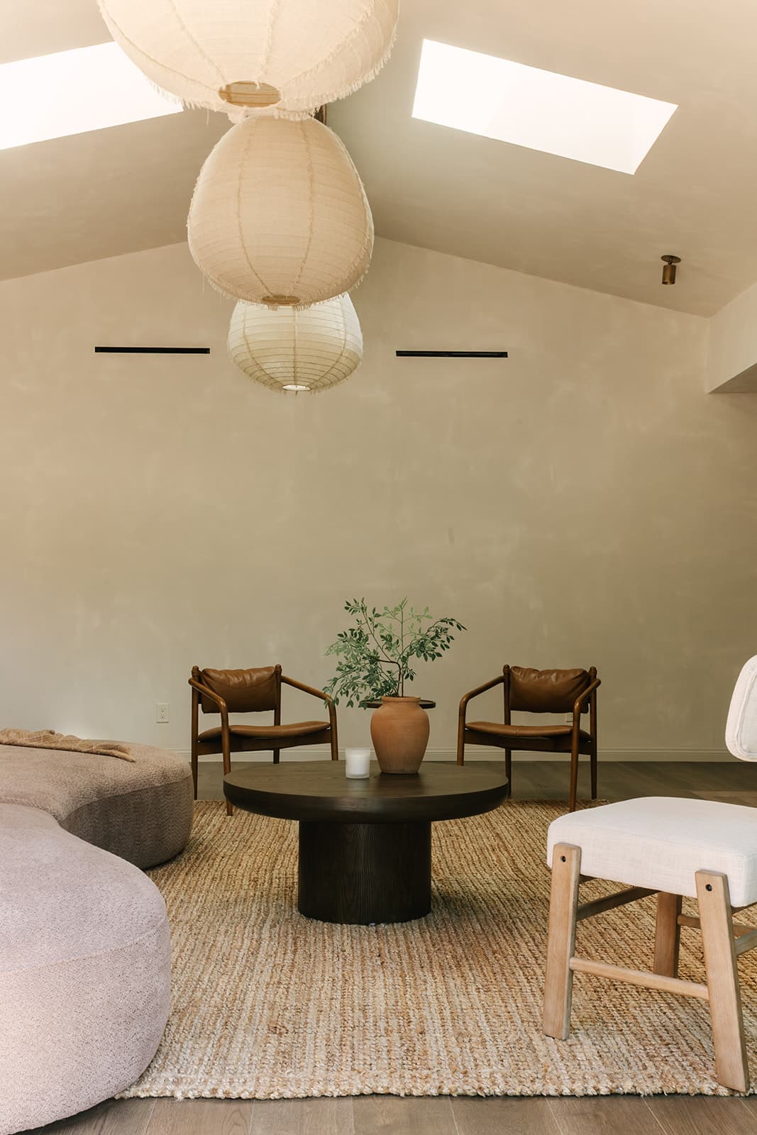 Minimalist living room with two brown leather chairs, a round dark wood coffee table holding a terracotta vase with green foliage, a light beige rug, and a soft curved sofa under white pendant paper lanterns.