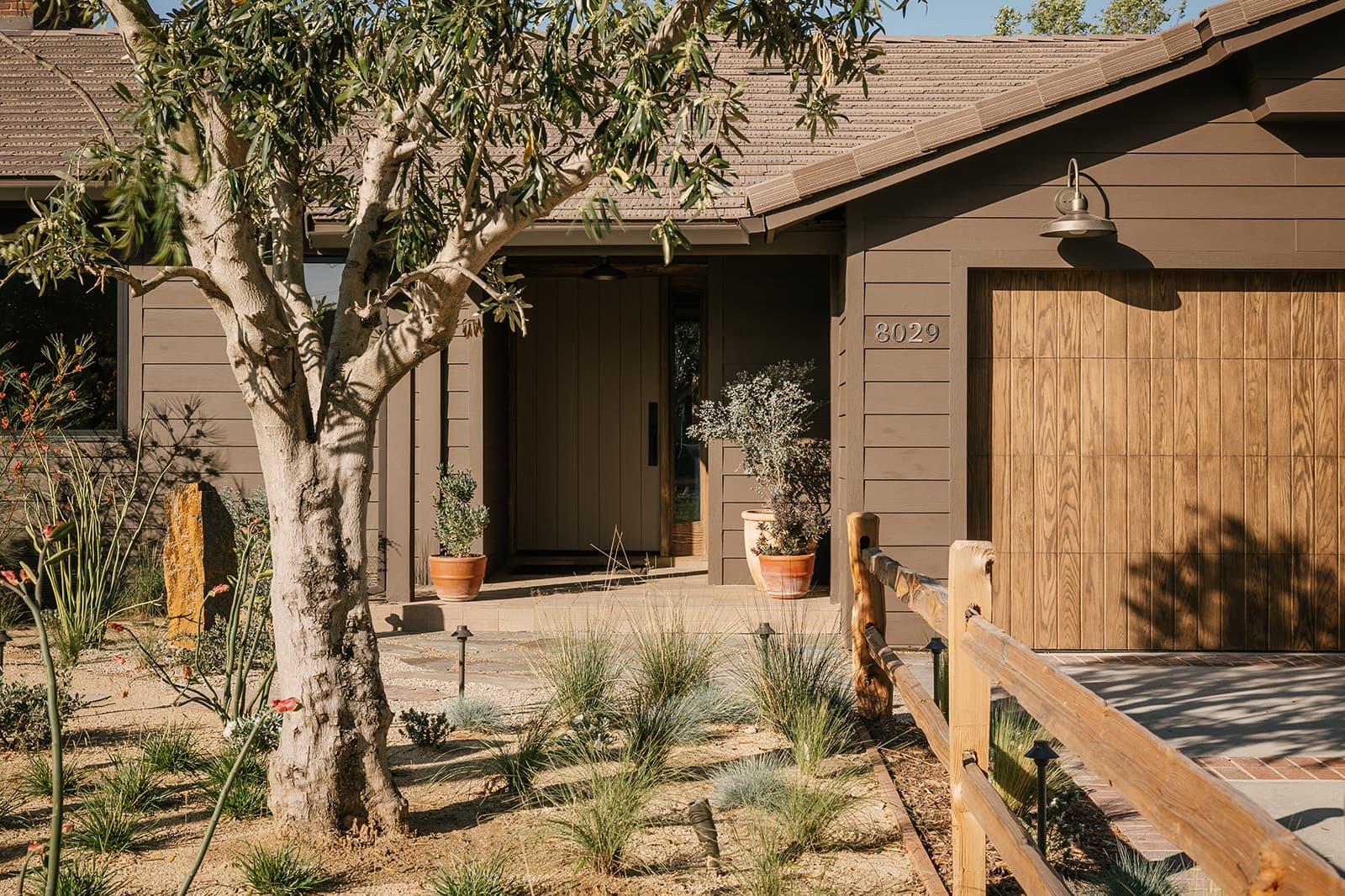 Front entrance of a modern brown house with wooden garage door, potted plants, and a wooden fence along the driveway.