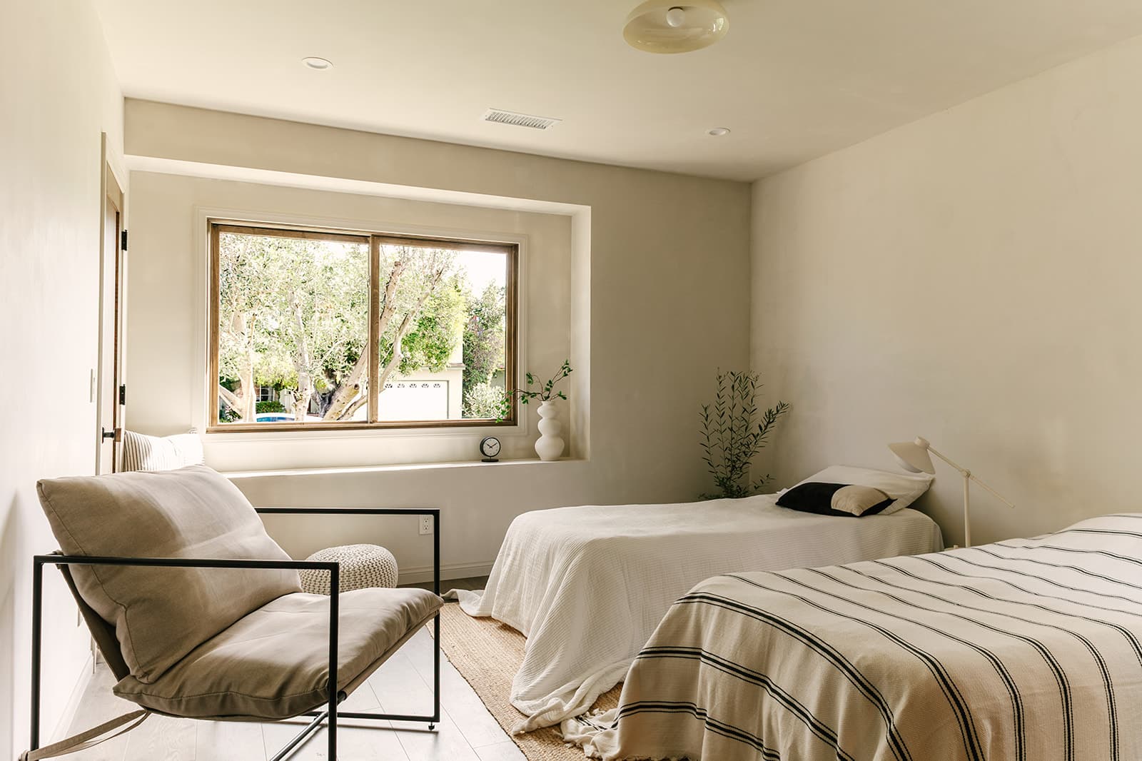 Minimalist bedroom with two single beds, beige and white linens, a beige cushioned chair, window with natural light, and small plants.