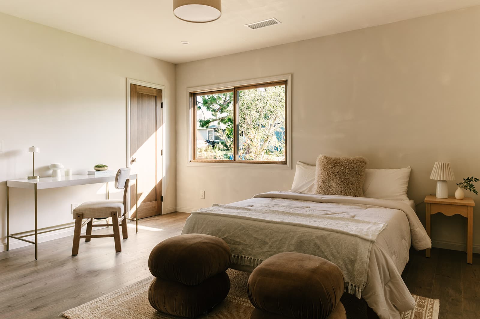 Minimalist bedroom with beige walls, a bed with white linens and a beige furry pillow, a wooden nightstand with a lamp and small plant, a white desk with a chair, and two brown poufs on a rug near the bed.
