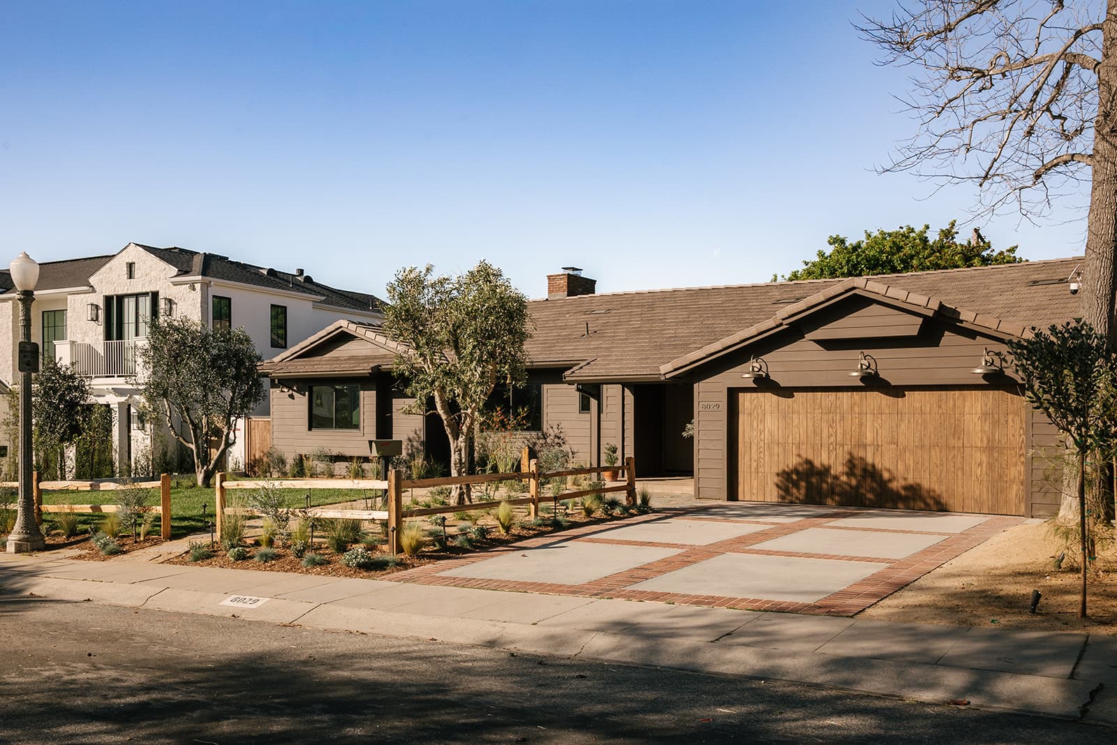 Single-story modern house with brown siding, wooden garage door, and a front yard with trees and a wooden fence.