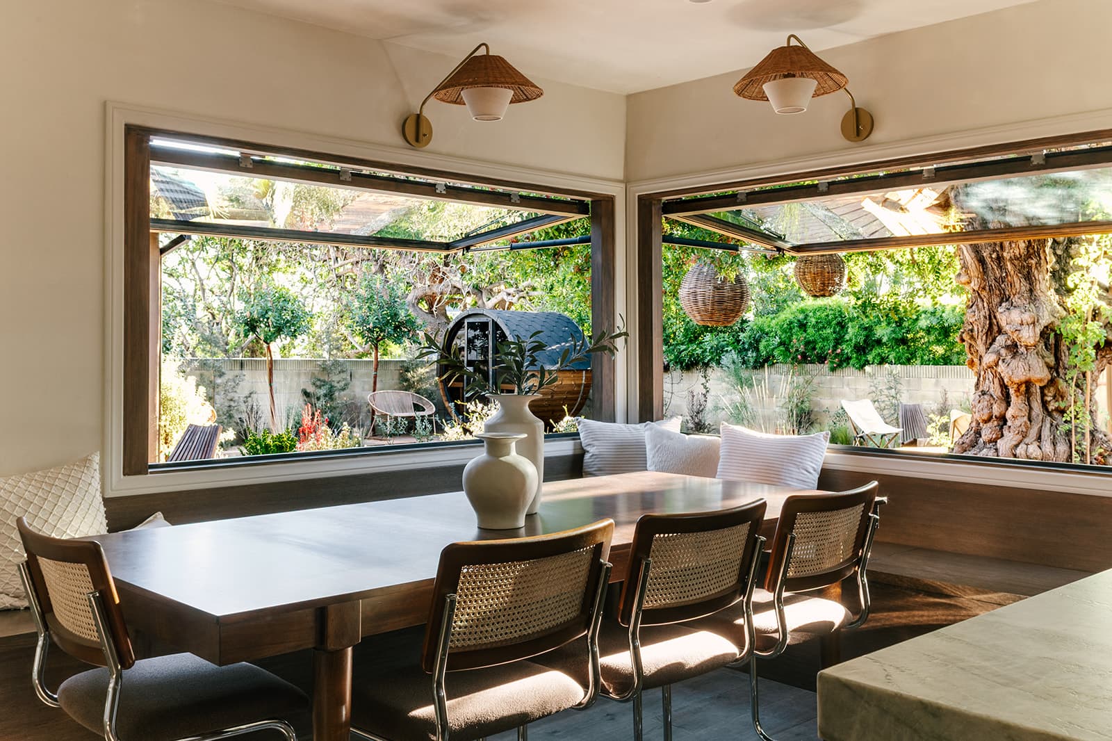 Sunlit dining area with wooden table, wicker chairs, vases with greenery, and large open windows showing a garden with trees and outdoor seating.