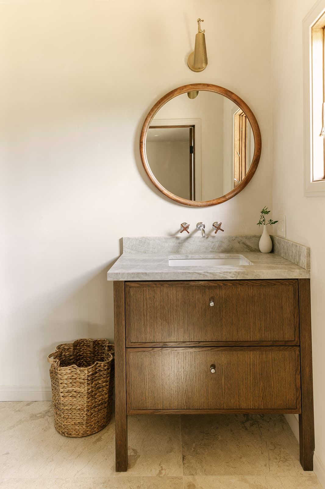 Minimalist bathroom vanity with marble countertop, round wooden mirror, brass wall light, and woven basket on tiled floor.