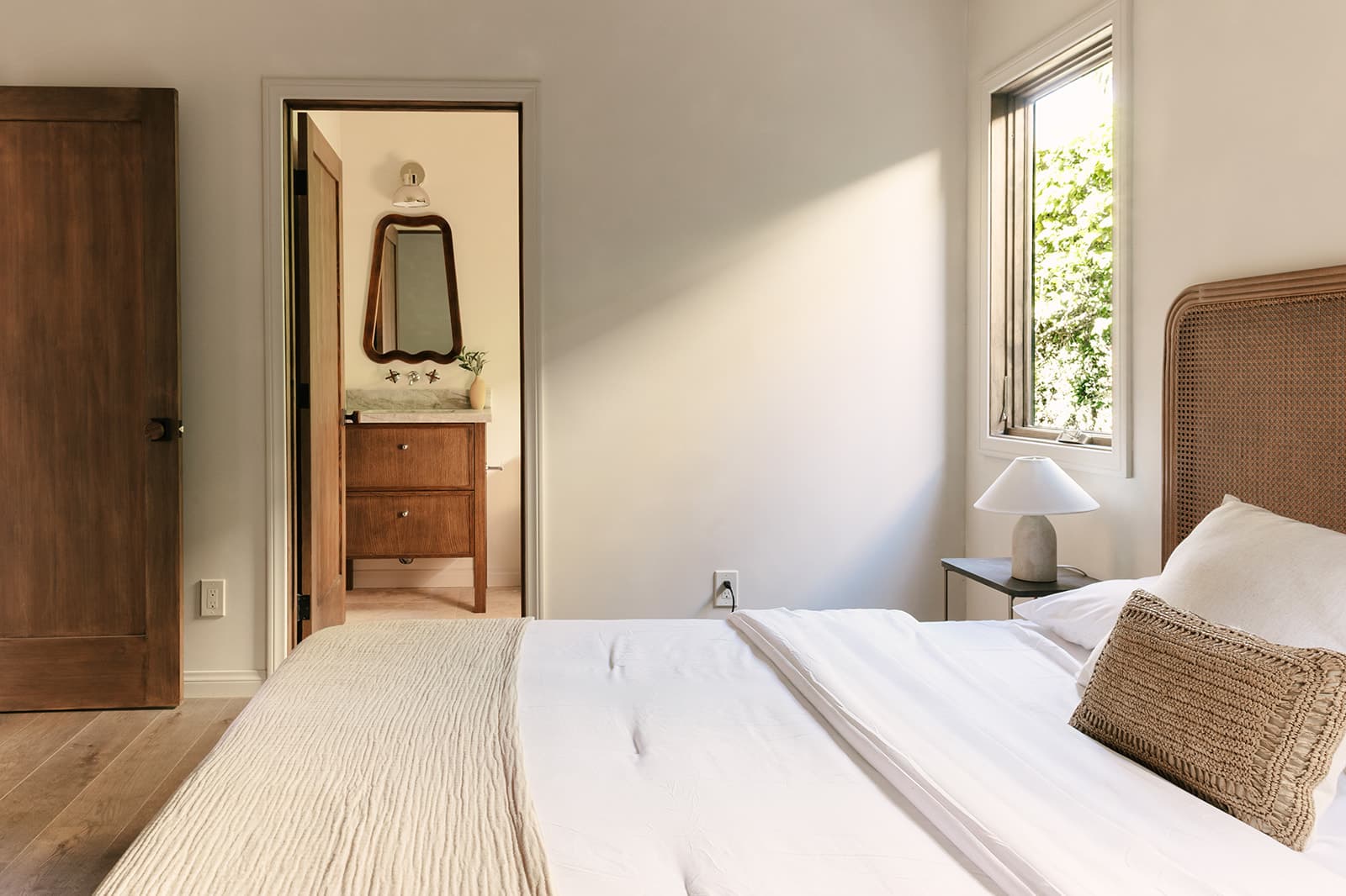 Minimalist bedroom with white bedding, a beige textured throw, a woven pillow, a wooden headboard, a bedside table with a white lamp, and an open door leading to a bathroom with a wooden vanity and a mirror.