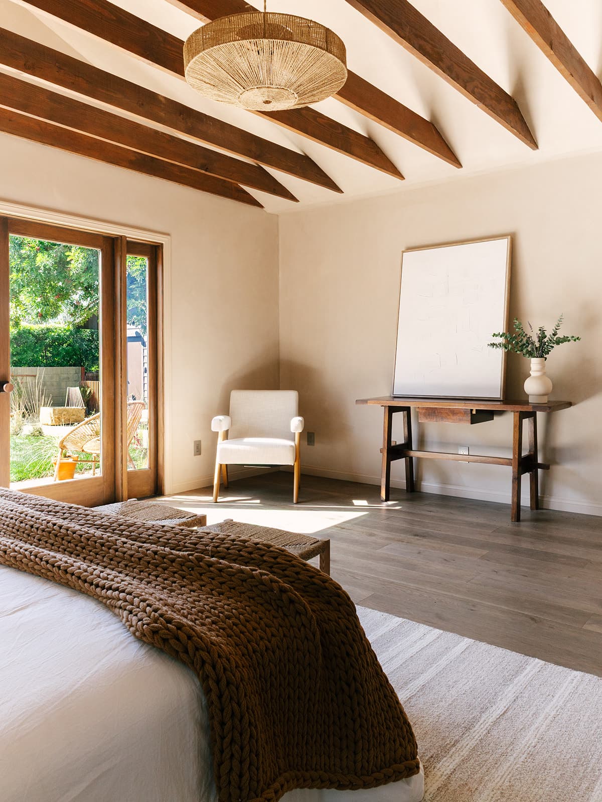 Bright bedroom with exposed wooden ceiling beams, a white armchair, wooden table with a blank frame and vase, and a bed with a chunky brown knitted blanket near glass doors opening to a garden.