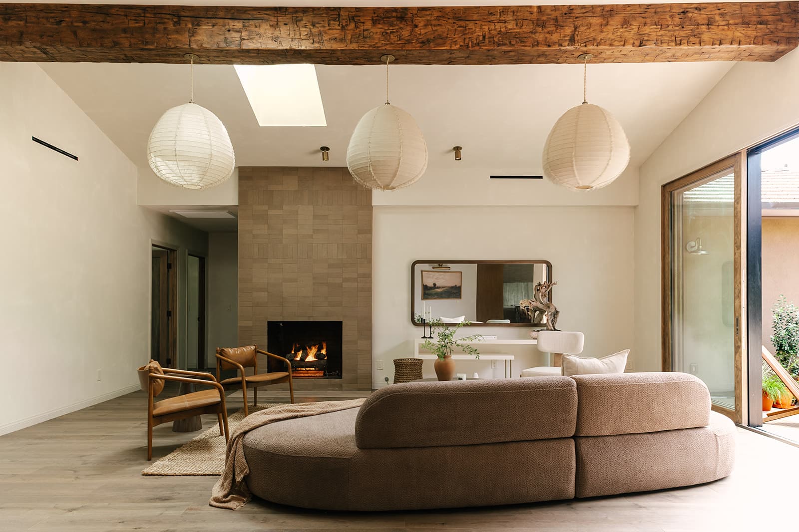 Cozy living room with large curved beige sofa, two leather armchairs, a fireplace, three hanging paper lanterns, and a glass door leading outside.