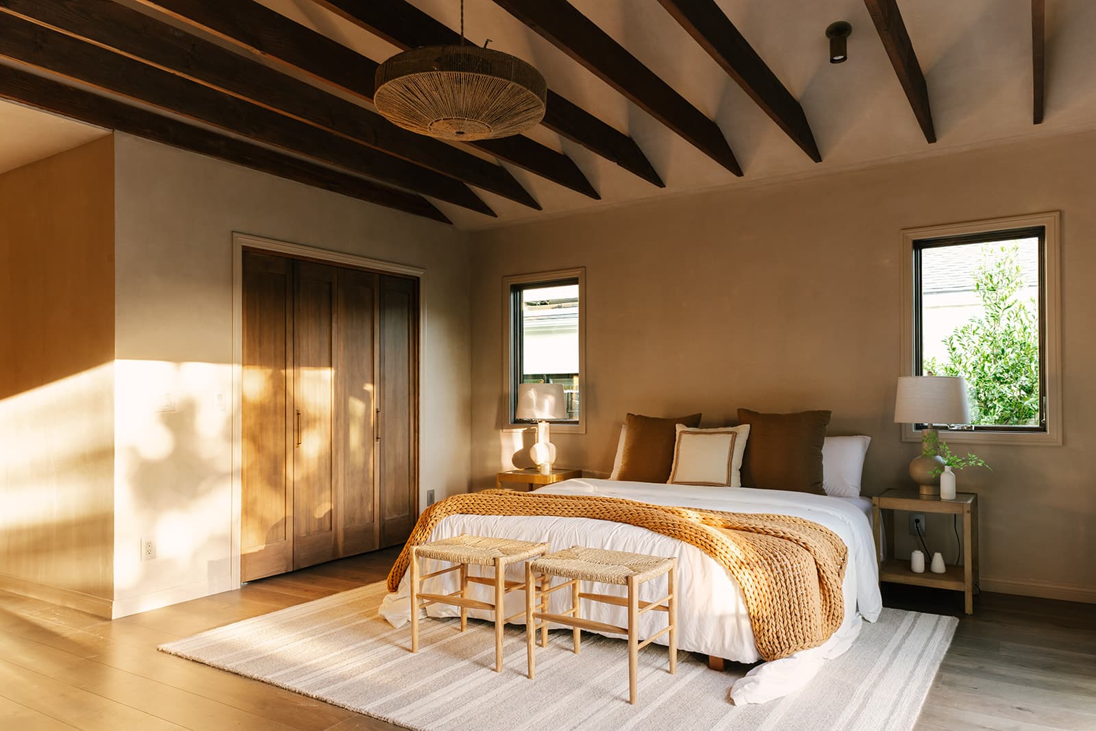 Bedroom with a white and beige bed, two wooden stools at foot, two windows, and wooden ceiling beams.