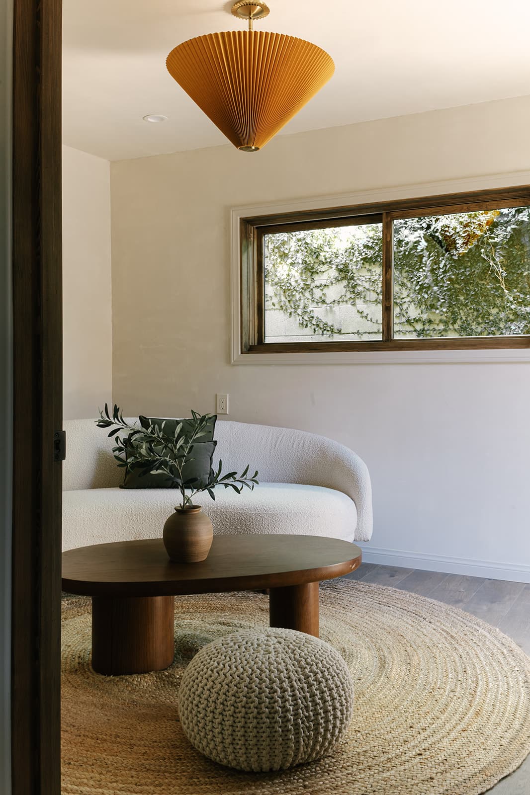 Minimalist living room with a white curved sofa, round wooden coffee table with a vase, knitted pouf, and a large window showing greenery outside.