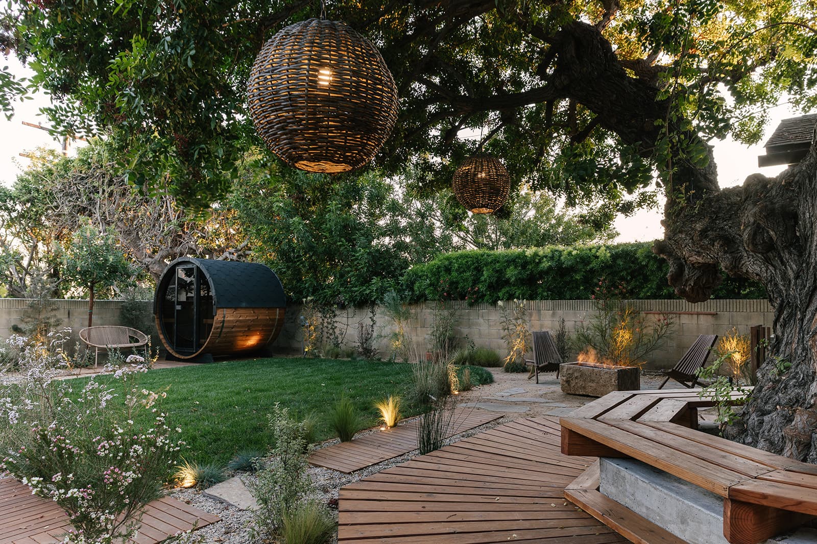 Cozy backyard with wooden deck seating around a large tree, hanging wicker lanterns, green lawn, fire pit with chairs, and a small barrel sauna near a concrete wall.