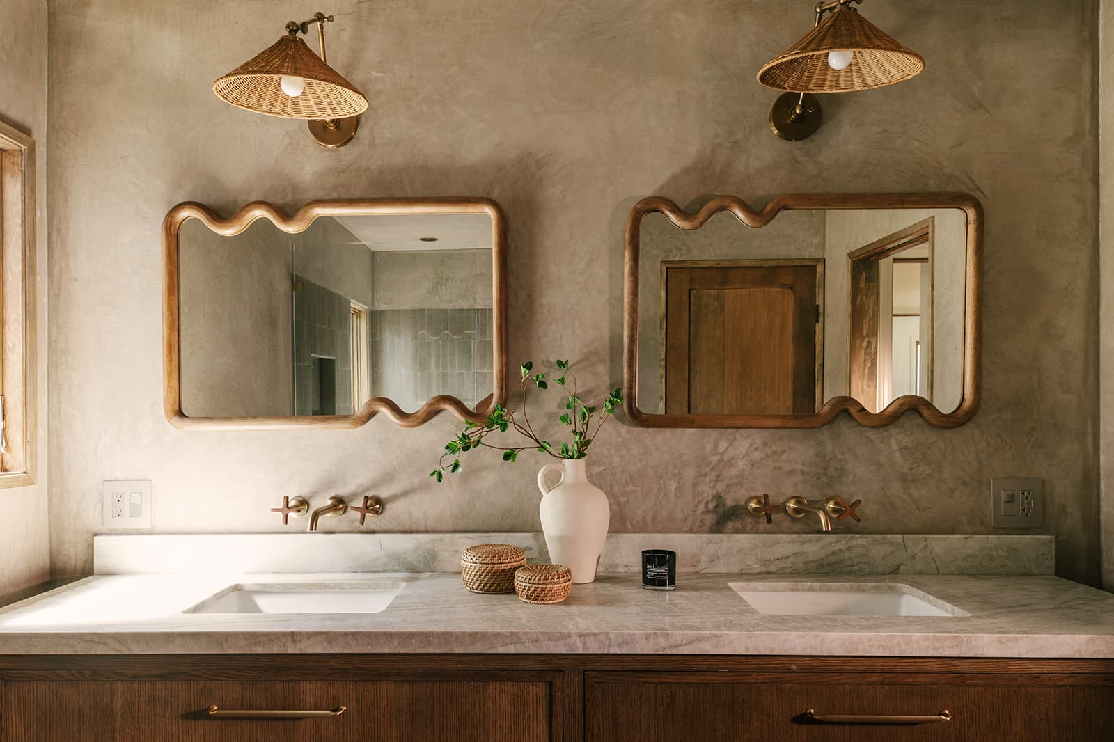 Modern bathroom countertop with two sinks, wavy-edged wooden mirrors, brass faucets, wicker light fixtures, and decorative vase with green branches.
