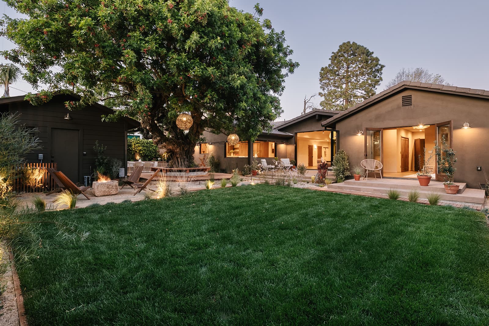 Backyard of a modern house with lush green lawn, large tree with hanging lights, stone fire pit, wooden chairs, and open doors leading indoors.