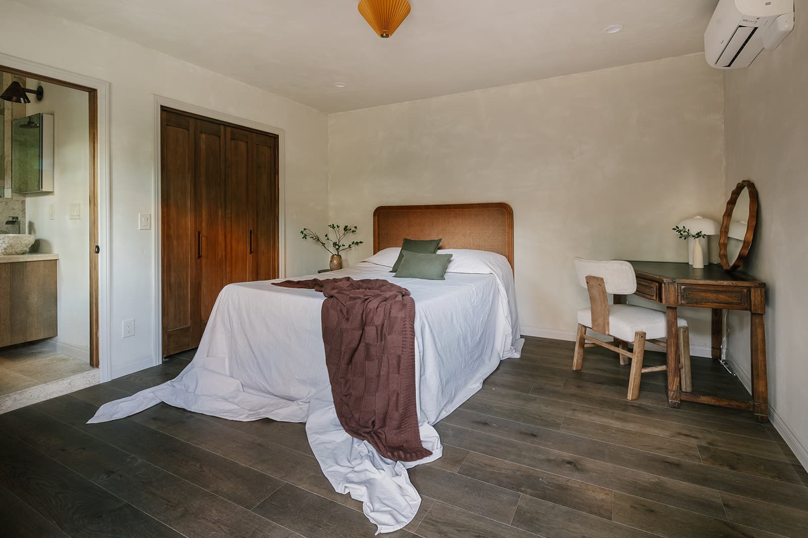 Minimalist bedroom with wooden floor, bed covered in white sheets and brown blanket, two green pillows, wooden closet, and wooden table with chair and round mirror.