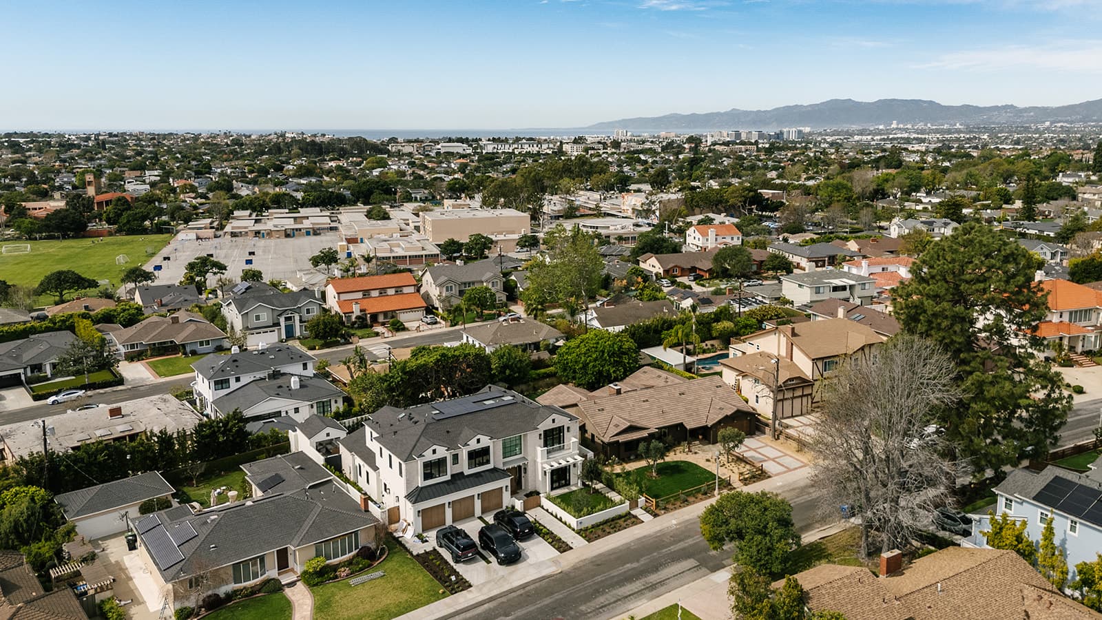 Aerial view of a suburban neighborhood with houses, green lawns, a school playground, and mountains in the distance under a partly cloudy sky.