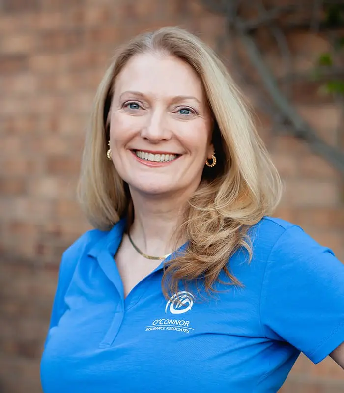 Smiling woman with long blonde hair wearing a blue O'Connor Insurance Associates polo shirt in front of a blurred brick wall.