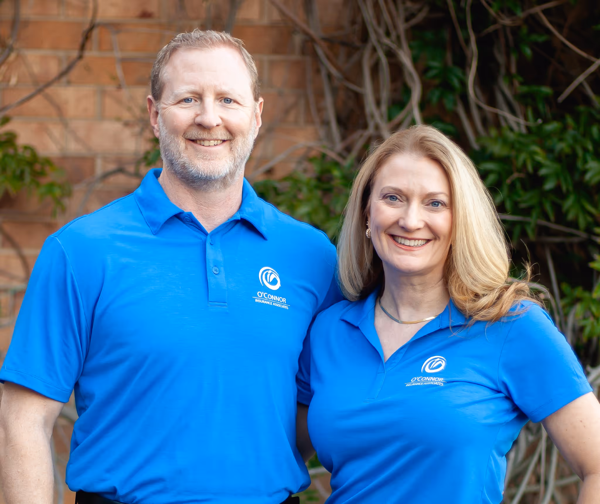 Smiling man and woman wearing matching blue O'Connor Insurance Associates polo shirts standing outdoors with a brick wall and greenery in the background.