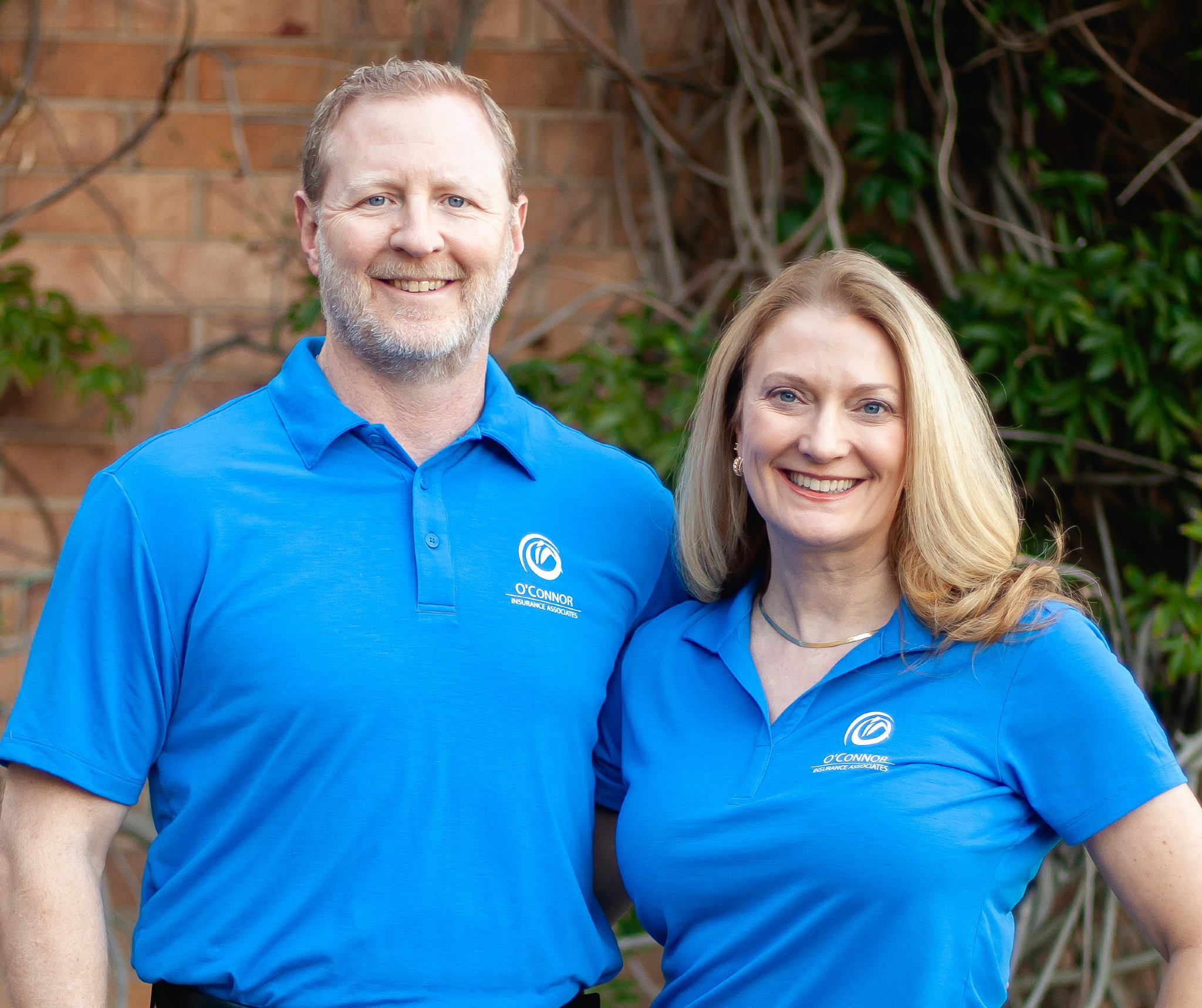 Smiling man and woman wearing matching blue O'Connor Insurance Associates polo shirts standing outdoors with a brick wall and greenery in the background.