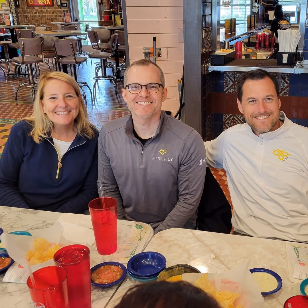Three people smiling and sitting closely together at a restaurant table with chips, salsa, and red drinks.