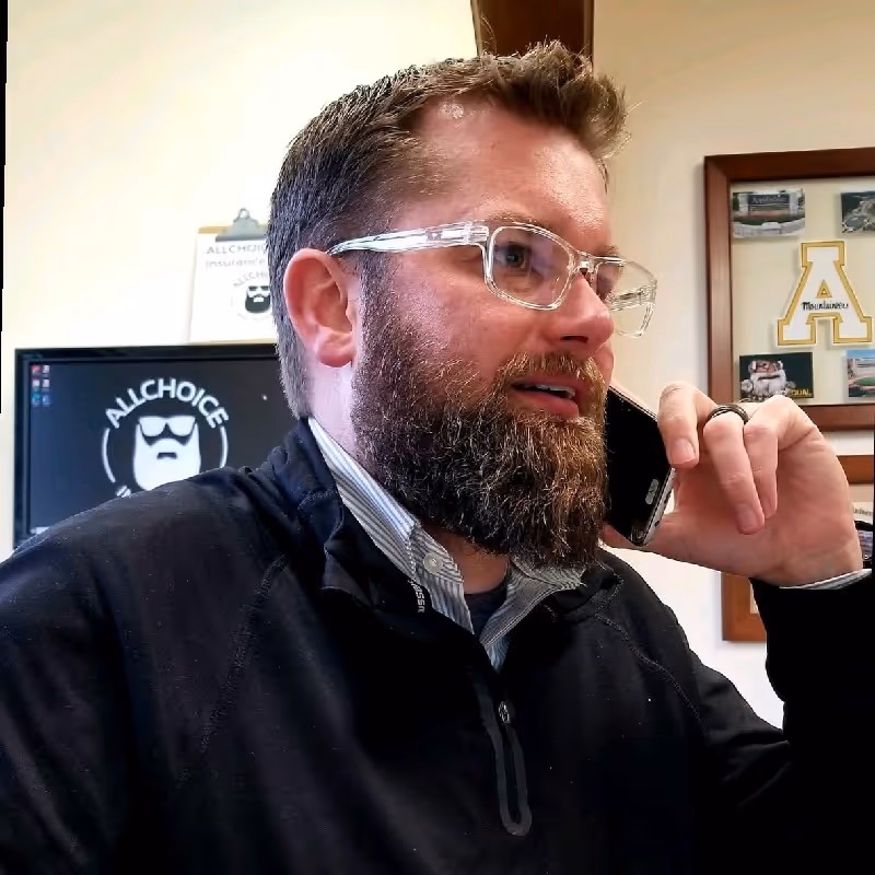 Photo of Jared Bellmund. He is wearing clear glasses and a black jacket talking on a smartphone in an office with framed pictures and a computer screen in the background.