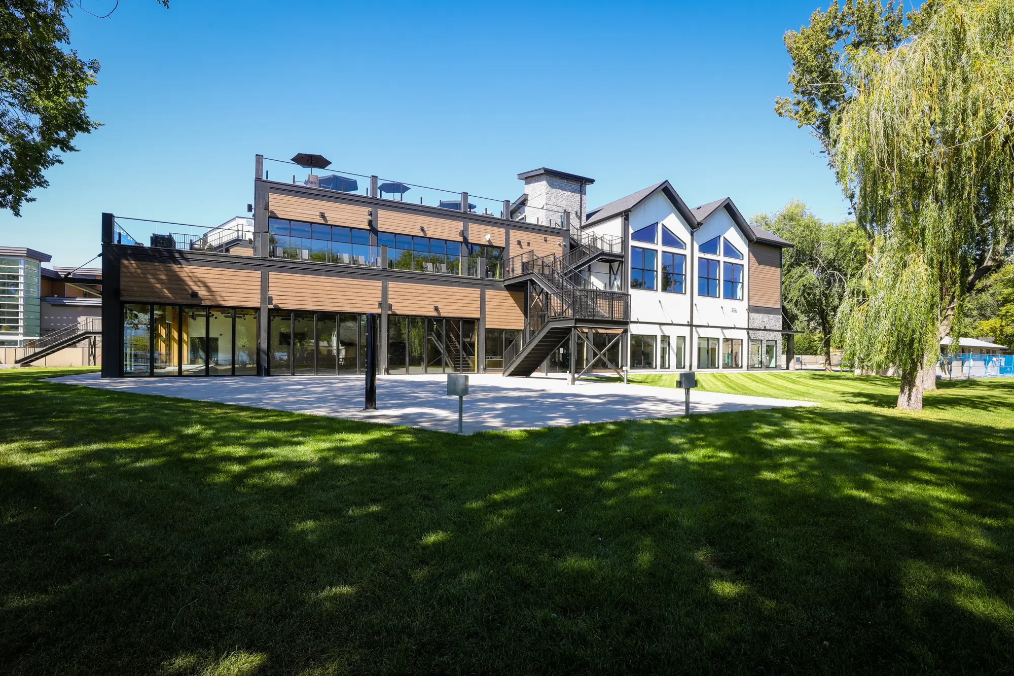 Modern two-story building with large windows, wooden paneling, outdoor stairs, and a rooftop patio overlooking a grassy lawn with trees.