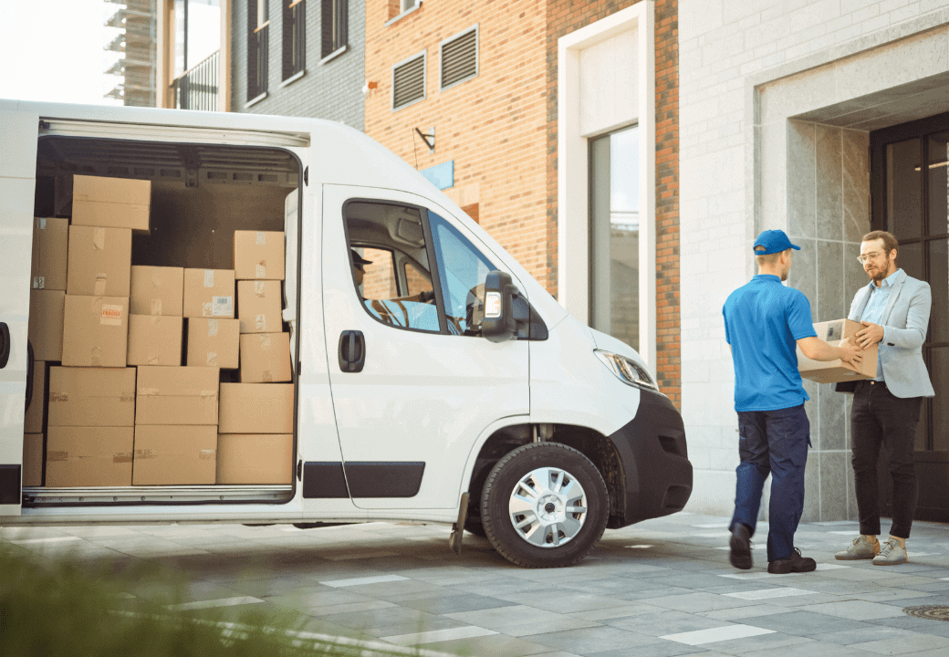 Van full of delivery boxes parked with door open and delivery man handing over a package to another man. 