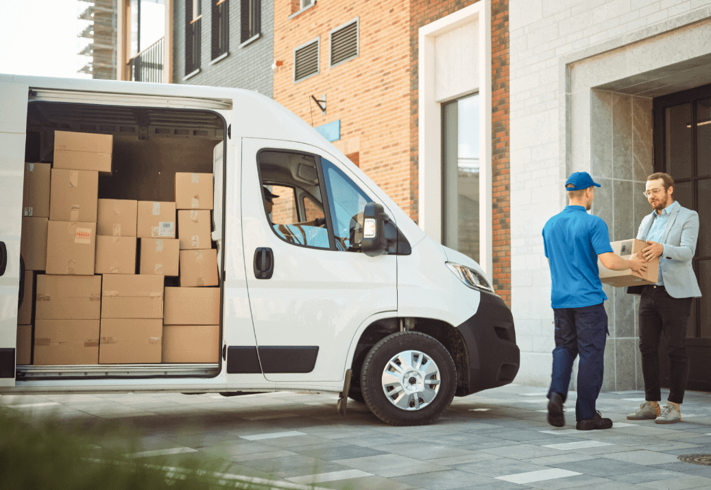 Van full of delivery boxes parked with door open and delivery man handing over a package to another man. 