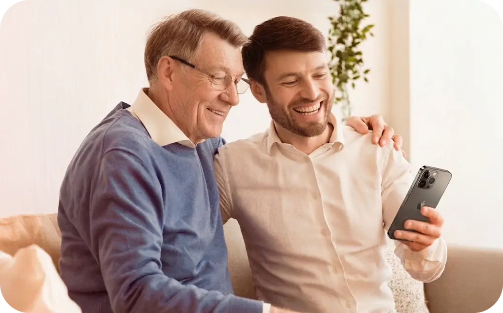 Älterer Mann und jüngerer Mann lachen, während sie auf einem Sofa ein Handy ansehen.