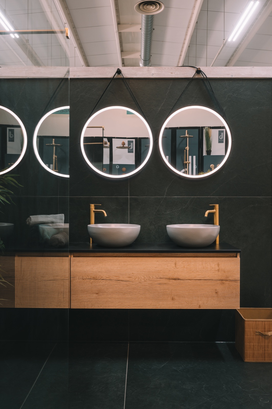 Twee waskommen met gouden kranen op een houten ribbelmeubel onder ronde spiegels met verlichting tegen een donkere wand.