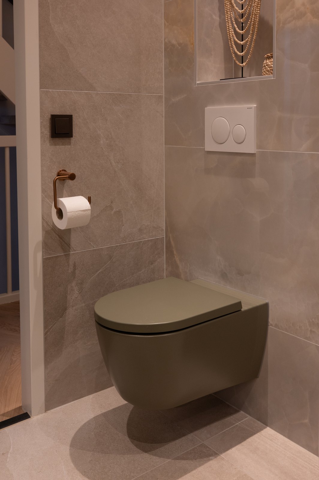 Olijfgroen wandcloset met bijpassende zitting tegen grijsbeige marmerlook-tegels, met koperen toiletrolhouder