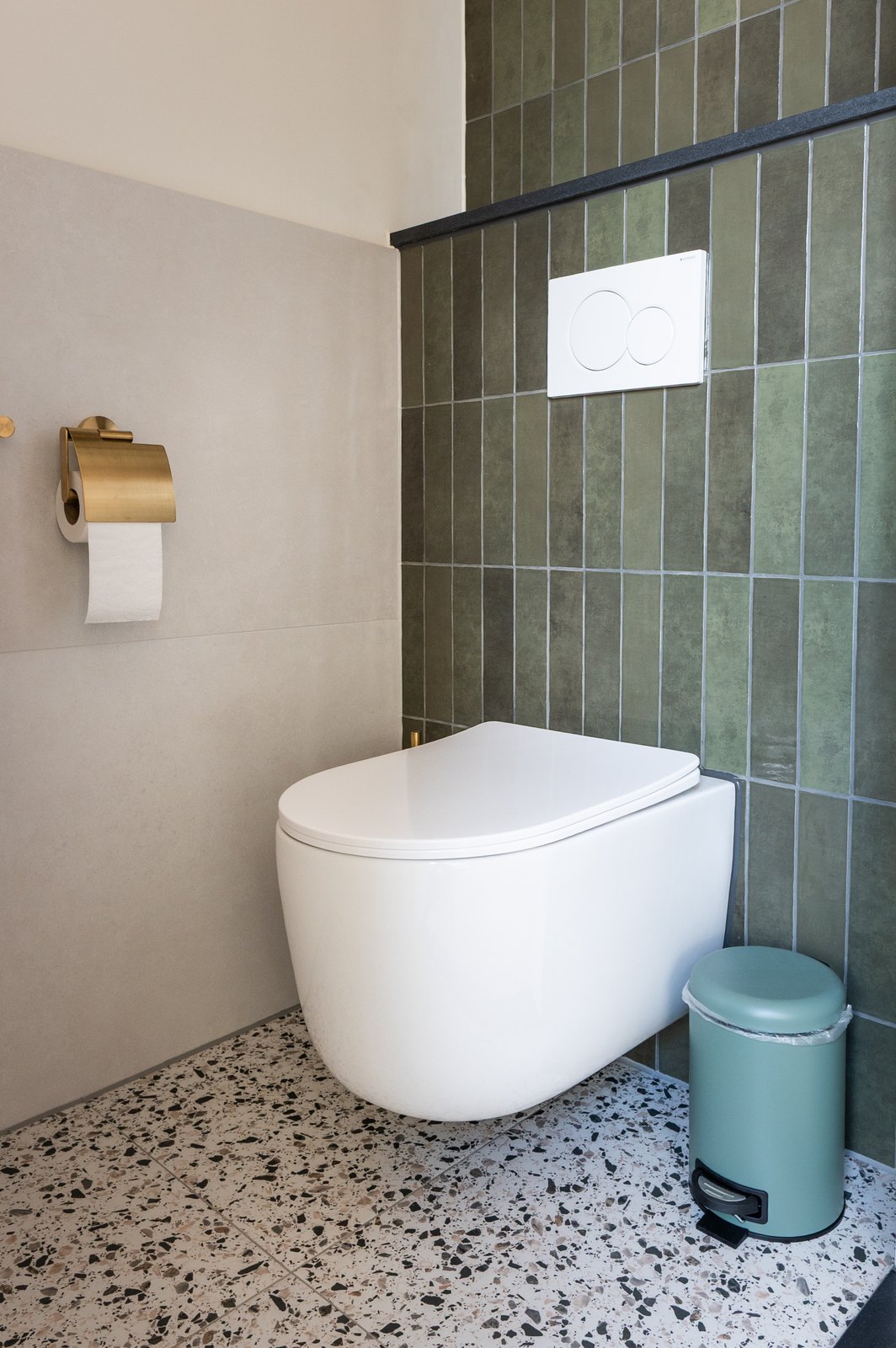 Toiletruimte met wit wandcloset tegen groene verticale tegels, terrazzovloer en een messing toiletrolhouder