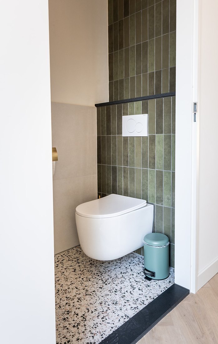 Wit wandcloset tegen groene staaftegels, met een terrazzovloer en een gouden toiletrolhouder