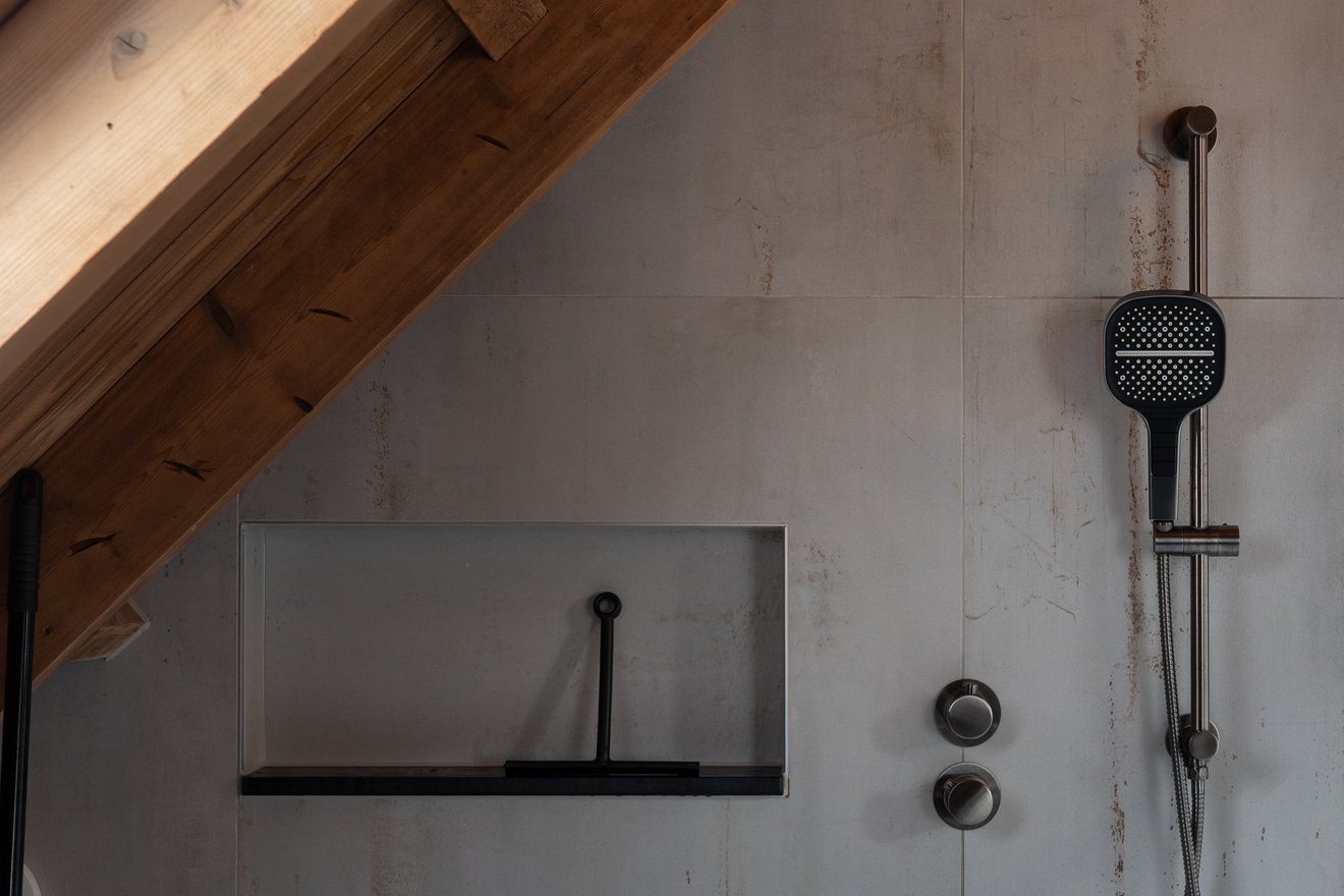 Inbouwnis en handdouche in een betonlook douchewand, naast een houten dakspant.