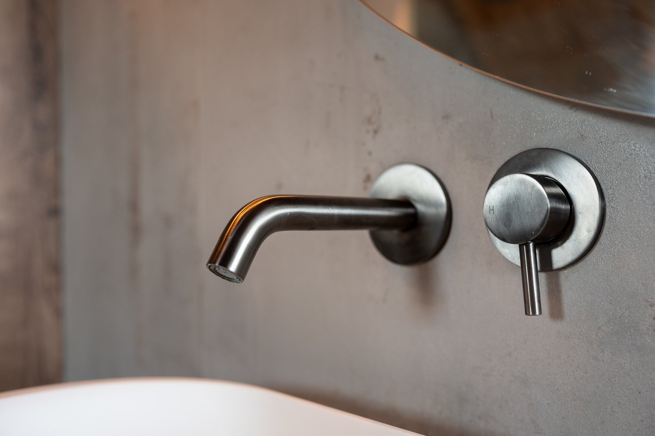 Wandkraan met uitloop en bedieningsknop in geborsteld metaal tegen een betonlook wand.