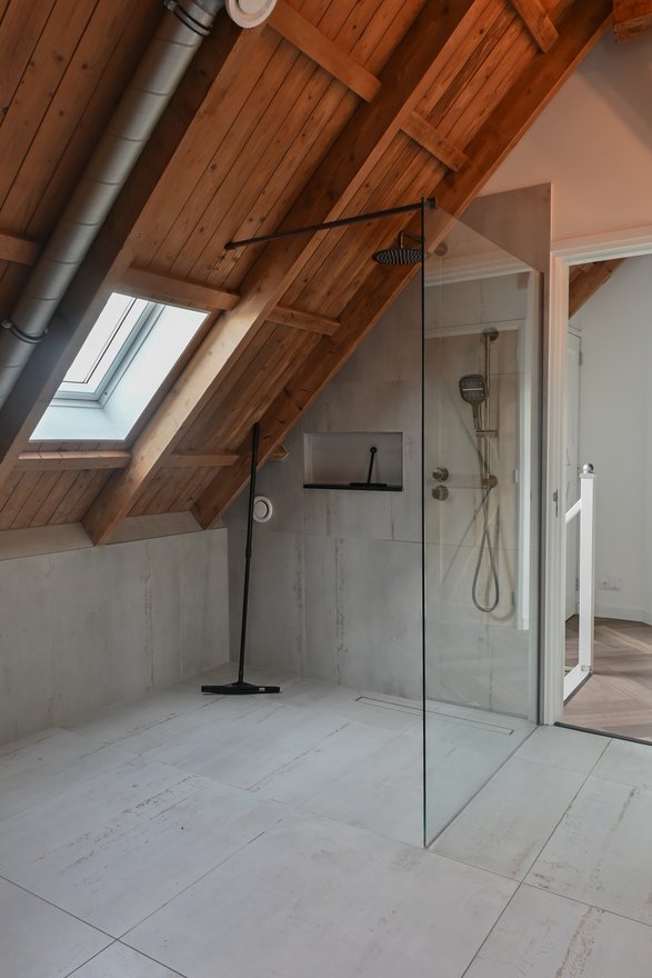 Zolderbadkamer met een inloopdouche achter glas, betonlook wanden en een houten dakconstructie met dakraam.