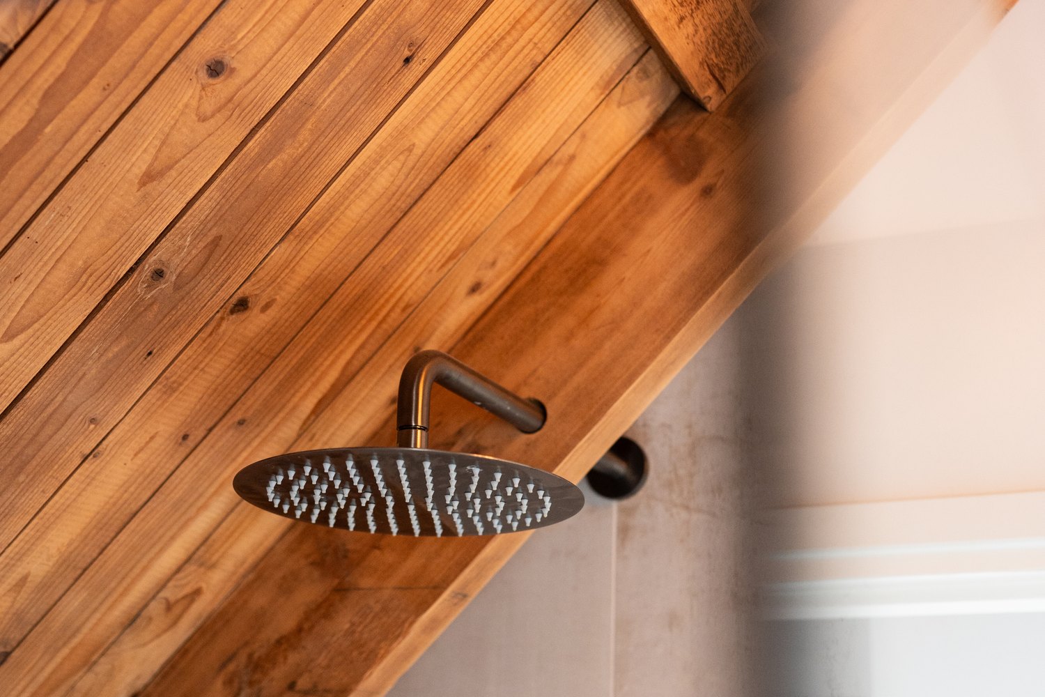 Ronde regendouchekop in donker metaal aan een gebogen arm, gemonteerd tegen het schuine houten dakbeschot van een zolderbadkamer, naast een wand in betonlook.