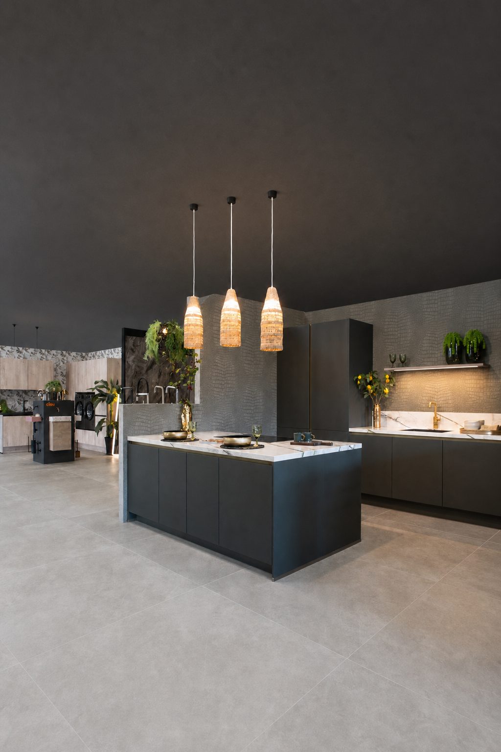 Antracietgrijs kookeiland in de Mega-showroom onder drie rotan hanglampen, met marmerlook werkblad, planten en een lichte tegelvloer onder een donker plafond