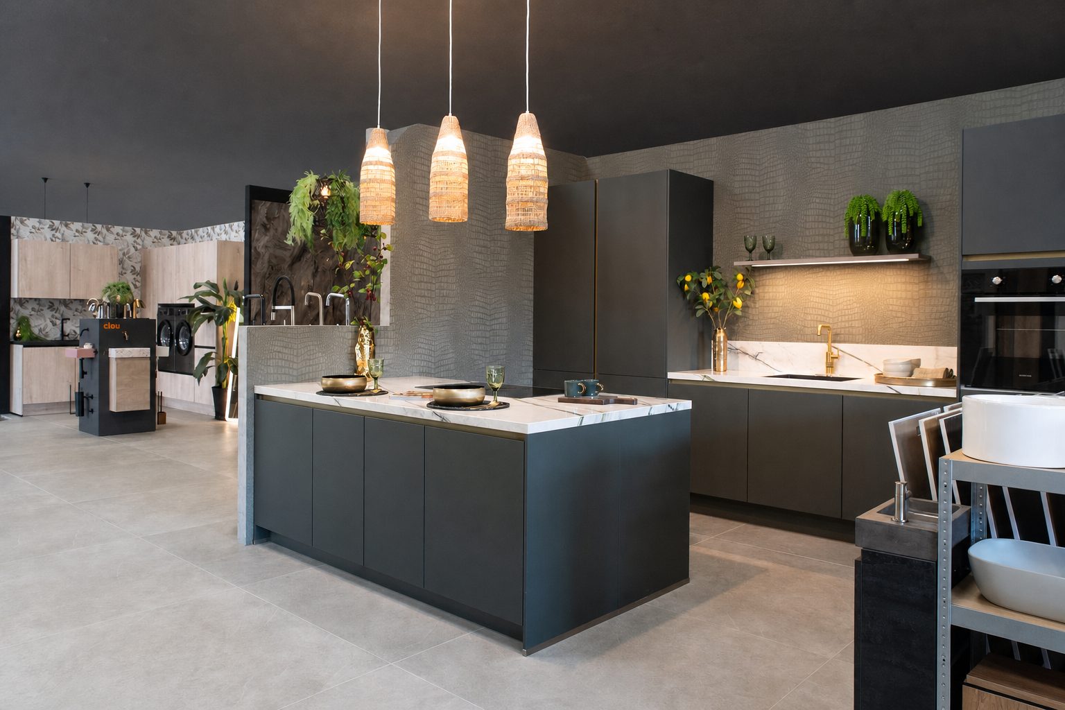 Ruime Mega-showroom met een antracietgrijs kookeiland onder drie rotan hanglampen, planten en glaswerk op het marmerlook werkblad en een lichte tegelvloer die doorloopt naar de andere opstellingen