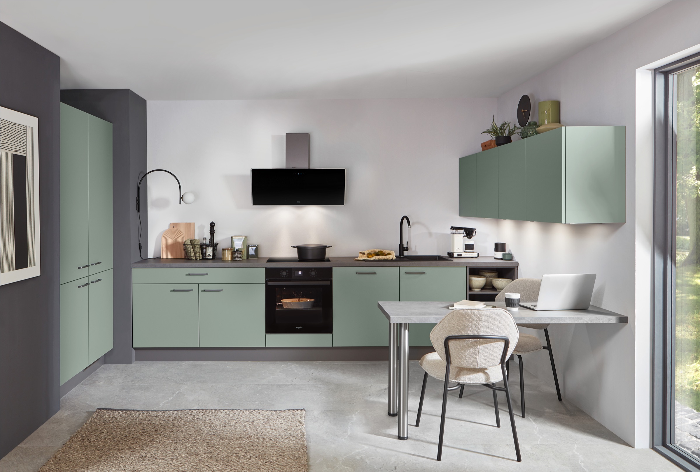 Moderne groene keuken in saliegroen met zwarte apparatuur en een kleine eethoek voor het raam