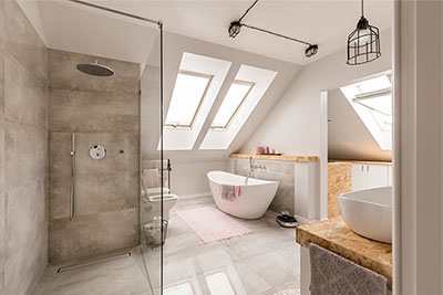 Zolderbadkamer met een betonlook douchewand en een vrijstaand bad onder twee dakramen