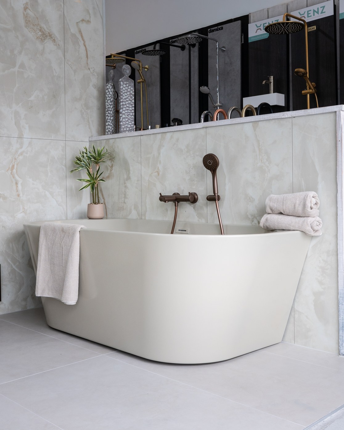 Vrijstaand bad met een bronzen badkraan en handdouche tegen een wand met marmerlook tegels in de showroom