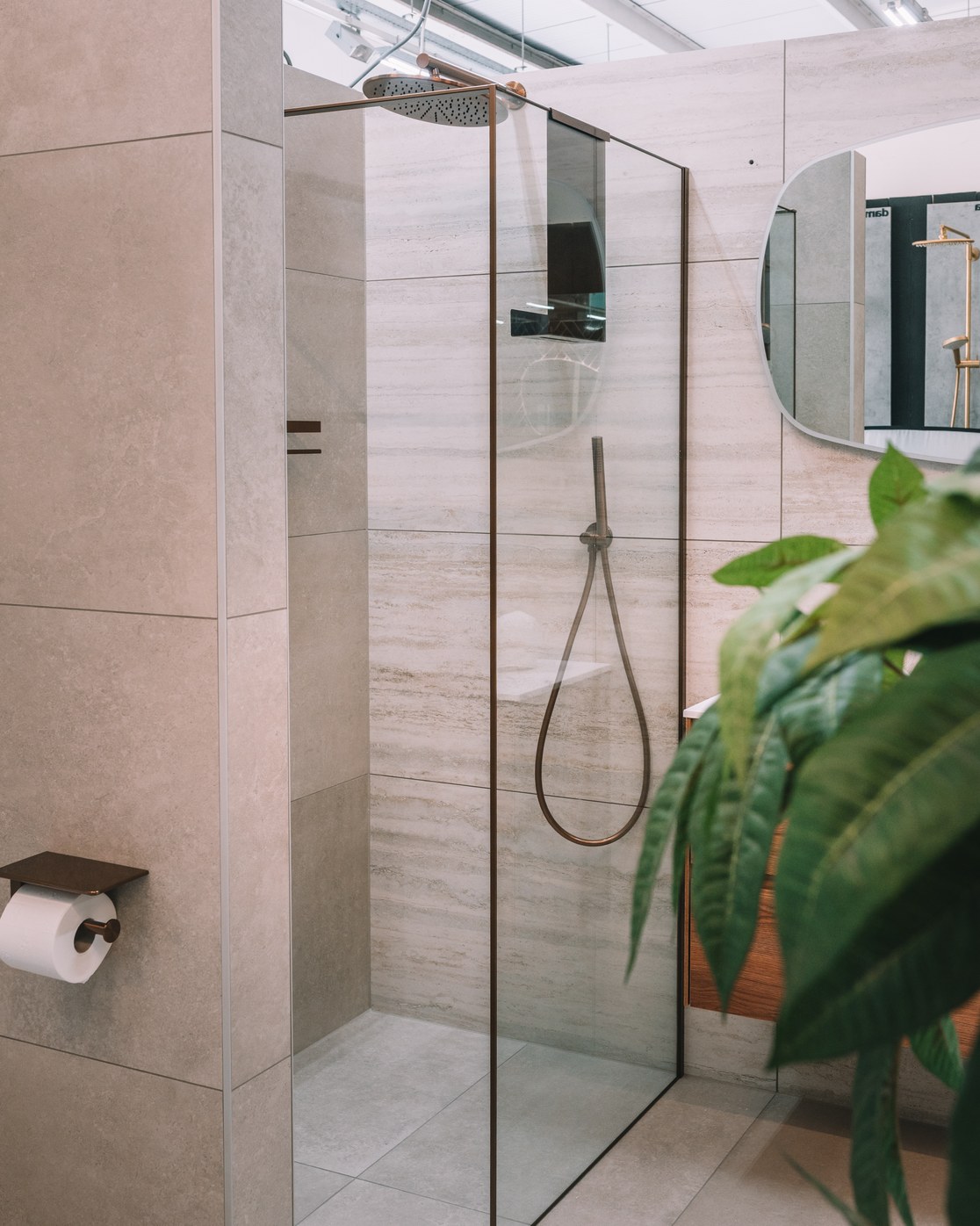 Inloopdouche met glazen wand en bronzen regendouche tegen travertijnlook tegels in de showroom
