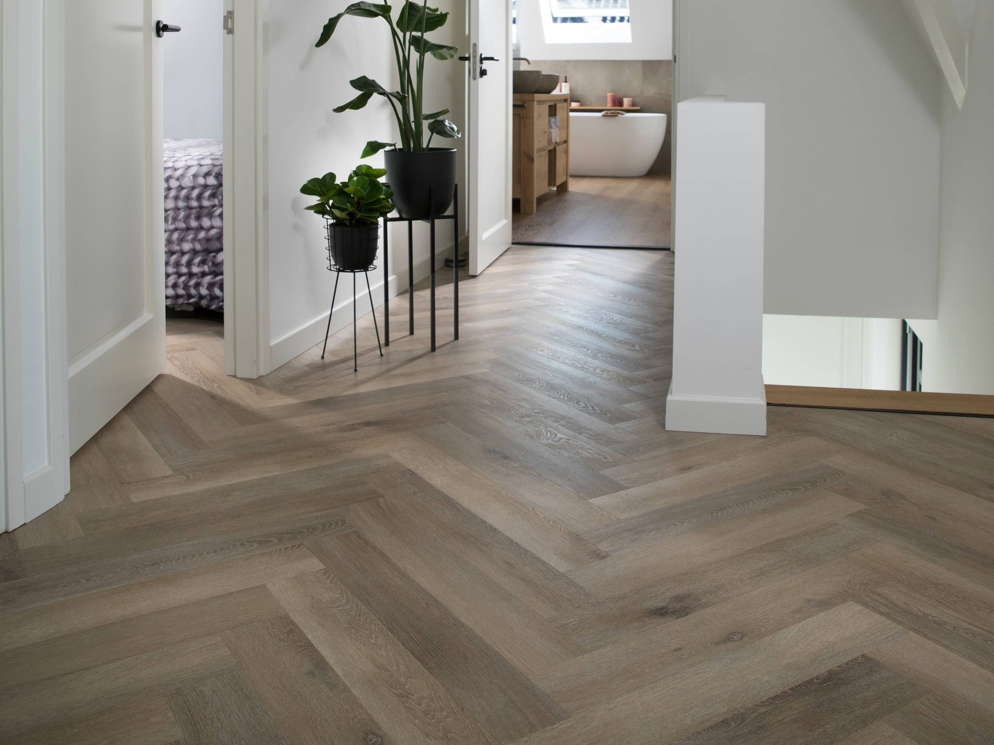 Overloop met een visgraatvloer, twee planten en doorkijk naar een slaapkamer en een badkamer